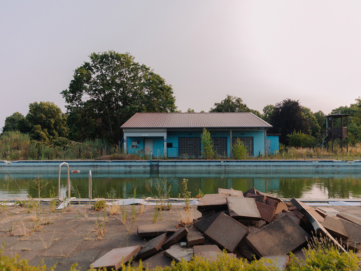 Blaues Pavillongebäude des Volkacher Freibads am Wasser, umgeben von Schilf und Bäumen im Abendlicht